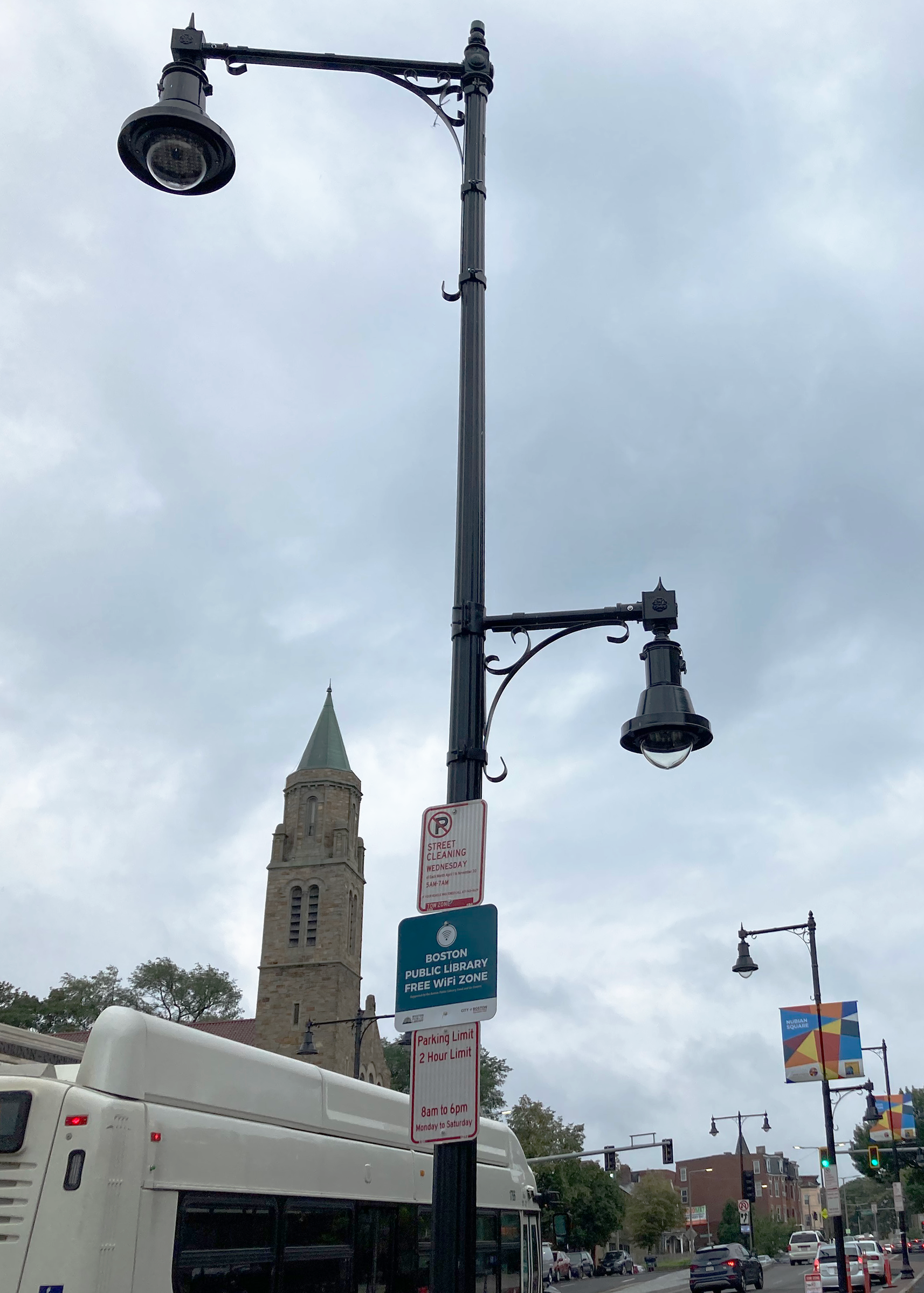 A pendant-style street light has one large, down-facing light hang approximately 4' over the roadway. A second, smaller pendant-style light  is on the same pole, but directed over the sidewalk. 
